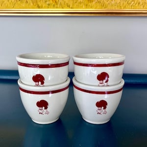 Może przedstawiać: Cztery białe ceramiczne miski z czerwonym paskiem na obrzeżu i ilustracją postaci z kreskówki. Miski są ułożone parami, prezentując wzór. Postać z kreskówki jest czerwona i wygląda na szefa kuchni.