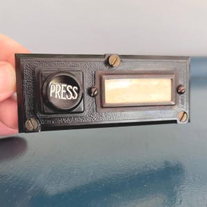 May include: A rectangular black doorbell button with a textured finish, featuring a circular push button labelled with the word PRESS in white lettering. To the right is a rectangular nameplate slot with a blank insert, secured by four screws.