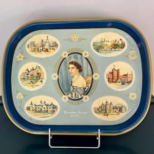 Op de afbeelding: Vintage blauw en goud Coronation Souvenir dienblad uit juni 1953, met een portret van koningin Elizabeth II en illustraties van verschillende gebouwen. De lade heeft een rechthoekige vorm met afgeronde hoeken en een metalen standaard.
