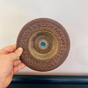 Könnte beinhalten: Eine braune Keramikschale mit einem strukturierten, gemusterten Rand und einem zentralen türkisfarbenen Akzent. Die Schale hat ein kreisförmiges Design mit konzentrischen Ringen und einer leicht vertieften Mitte. Die Schale wird von einer Hand gehalten.