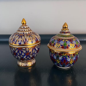 May include: Two small, ornate, lidded ceramic jars with gold trim and intricate blue, white, red, and green floral patterns. The jars are on a dark blue surface.