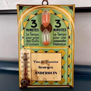 Könnte beinhalten: Vintage Wanduhr mit einer 3-Minuten-Sanduhr und einem Thermometer. Der Timer ist mit grünen und goldenen Akzenten verziert und enthält Text auf Französisch. Der untere Teil trägt die Aufschrift "Vins de Touraine Georges ANDEROUIN".