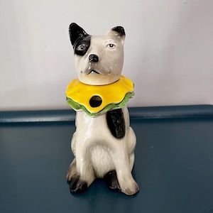 Könnte beinhalten: Keramikhundefigur mit gelb-grünem Kragen. Der Hund ist weiß mit schwarzen Flecken und einem schwarzen Ohr. Die Figur sitzt aufrecht und hat ein skurriles Design. Der Kragen des Hundes hat ein Loch in der Mitte.