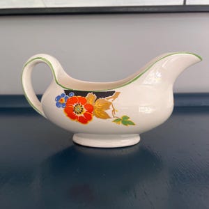 Puede incluir: Una salsera de cerámica blanca con un borde verde y un diseño floral. El diseño presenta una gran flor naranja con detalles azules y hojas verdes.