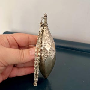 Peut inclure: Un pendentif argenté en forme de goutte d'eau avec des motifs géométriques et floraux complexes. Le pendentif est suspendu à une chaîne délicate. Le design présente un dessus décoratif et un fond pointu. Le pendentif est tenu dans une main.