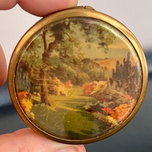Peut inclure: Un poudrier rond en métal doré avec une illustration de paysage colorée et brillante sur le couvercle. L'image représente une scène de jardin luxuriante avec des arbres verts, des fleurs orange et jaunes vives et un sentier au loin.