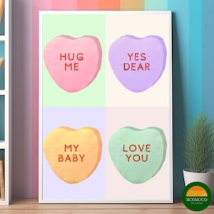 May include: A framed print showing four pastel-coloured conversation heart sweets. Each heart has a different message: "HUG ME," "YES DEAR," "MY BABY," and "LOVE YOU." The artwork is displayed on a wooden surface.