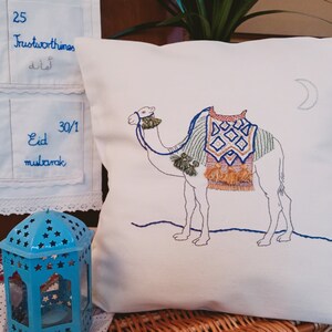 May include: White pillow with embroidered camel with a colorful saddle and a crescent moon in the top right corner. The text "Eid Mubarak" is embroidered on a white pillowcase behind the pillow.
