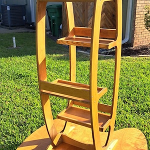 Può includere: Una libreria alta in legno con un design unico a forma di tavola da surf. I ripiani marrone chiaro sono sostenuti da una struttura curva e hanno una base ovale ampia. La libreria ha più ripiani per riporre e mostrare oggetti.