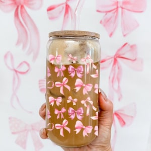 May include: A glass reusable coffee cup with a bamboo lid and a pink bow pattern. The cup is filled with iced coffee and has a clear glass straw.