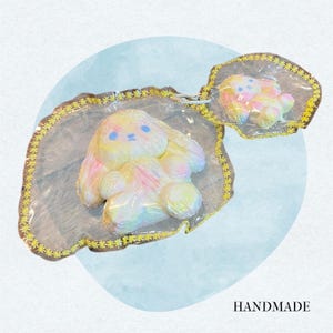 May include: A handmade, iridescent bunny-shaped item with blue eyes and pink and yellow accents, presented on a clear, star-edged tray. A smaller version is also visible. The word "HANDMADE" is at the bottom.