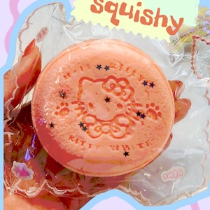 May include: A round, peach-colored product with a Kitty White design, surrounded by stars and paw prints, is visible through clear packaging. The text "So squishy" is in a yellow bubble. The product is on a light pink and blue background.