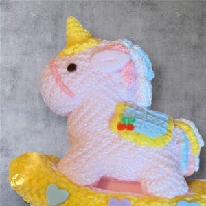 May include: A pink rocking horse figurine with a yellow horn and saddle. The horse has a light pink body with a textured surface, a yellow saddle, and a cherry design. The mane and tail are a mix of pink and blue.