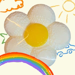 May include: A white, flower-shaped object with a yellow center, resembling a fried egg. The image has a playful, cartoonish style, with a rainbow, sun, cloud, and car doodles in the background. The flower has a textured surface.