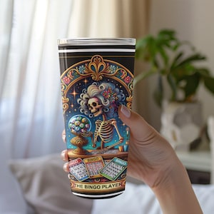 May include: A black and gold tumbler with a colorful illustration of a skeleton holding a bingo ball cage. The text "The Bingo Player" is at the bottom of the tumbler.
