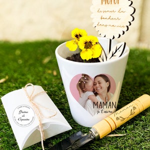 Peut inclure: Un pot de fleurs en céramique blanche avec une photo d'une mère et d'un enfant, des fleurs jaunes et un panneau en bois avec l'inscription "Merci". Une petite truelle et un sachet de graines sont également présents, le tout sur de l'herbe verte.