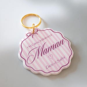 Op de afbeelding: Een roze en wit gestreepte sleutelhanger met het woord "Maman" in cursief, een hartje en de namen "Lucie et Sabri". De sleutelhanger heeft een gouden ring en de tekst "On t'aime infiniment".