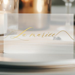 Könnte beinhalten: Eine durchsichtige Acryl-Platzkarte mit den Worten "La mariée" in goldener Kalligraphie.