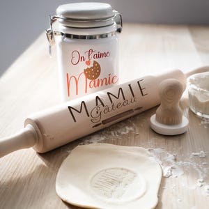 Peut inclure: Un pot en céramique blanche avec le texte "On l'aime Mamie" et un graphique de cookie. Un rouleau à pâtisserie en bois avec le texte "Mamie Gâteau" et un tampon avec un motif de cœur. Un petit bol en verre avec de la farine.