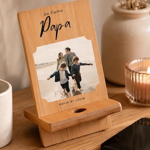 Peut inclure: Un support de téléphone en bois avec une photo de famille. Le support porte les mots "On t'aime Papa" et les noms "David et Lucie". Un smartphone noir est posé sur la table en bois.