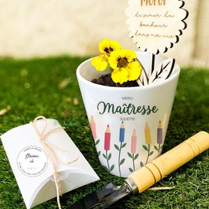 Peut inclure: Un pot en céramique blanc avec le mot "Maîtresse" et des illustrations de crayons, contenant des fleurs jaunes. Un panneau en bois avec le mot "Merci" est dans le pot. Une petite truelle et une boîte cadeau avec du texte sont également présents.