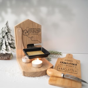 Könnte beinhalten: Ein Raclette-Grill aus Holz mit einer schwarzen Pfanne, die geschmolzenen Käse enthält, beleuchtet von zwei Kerzen. Ein kleines Holzschneidebrett mit einem Käsemesser und dem Text "La planche a charcuterie c'est le tapis rouge de la raclette" ist ebenfalls vorhanden. Ein kleiner dekorativer Weihnachtsbaum befindet sich im Hintergrund.