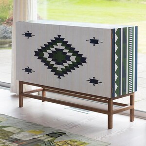May include: A white cabinet with a geometric pattern in green, blue, and white. The cabinet has two doors and a wooden base.
