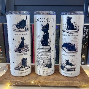 Peut inclure: Trois gobelets isothermes blancs avec des illustrations de chats noirs et du texte. L'un dit "Bookish Things" avec un chat sur des livres. Les autres disent "new book smell", "endless TBR", "cozy blanket", "emotionally attached", "one more chapter", et "comfy couch".