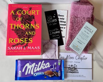 May include: A gift box containing a book titled "A Court of Thorns and Roses", a Milka Oreo chocolate bar, pink gloves, bookmarks, a wax melt, a tea bag, and a card with the text "One More Chapter". The book is by Sarah J. Maas.