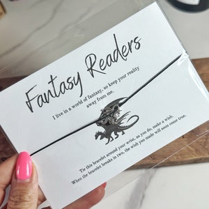 Op de afbeelding: Een zwart koordarmband met een zilveren drakenbedel. De kaart die aan het armband is bevestigd, heeft de tekst "Fantasy Readers" en bevat de tekst "Ik leef in een wereld van fantasie, dus houd je realiteit van me af."  "Bind dit armbandje om je pols, en doe daarbij een wens. Wanneer het armbandje in tweeën breekt, zal de wens die je hebt gedaan, snel uitkomen."
