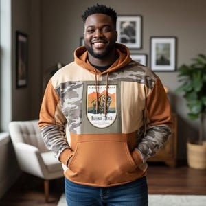May include: A hooded sweatshirt with a color block design featuring shades of tan, brown, and camouflage print. The front of the sweatshirt has a patch with the text "Buffalo Trace" and an image of a bison. The person is wearing blue jeans.