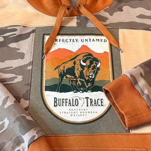 May include: A patchwork hoodie with a large graphic featuring a bison and the words "Buffalo Trace Kentucky Straight Bourbon Whiskey." The hoodie has a camouflage pattern on the sleeves and a rust-colored hood and drawstring.