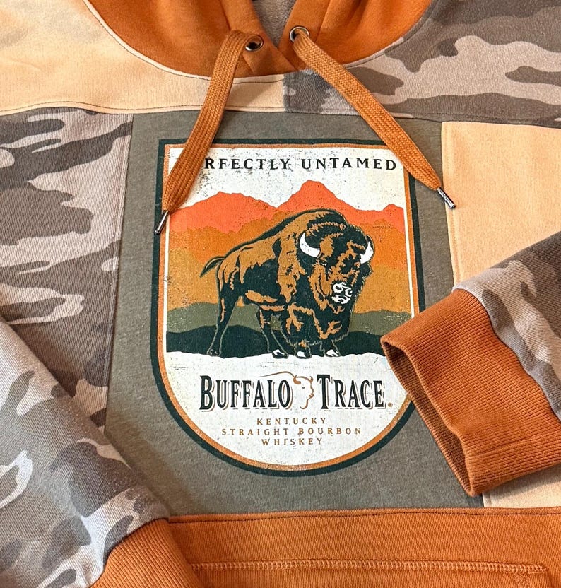 May include: A patchwork hoodie with a large graphic featuring a bison and the words "Buffalo Trace Kentucky Straight Bourbon Whiskey." The hoodie has a camouflage pattern on the sleeves and a rust-colored hood and drawstring.