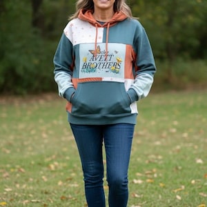 Könnte beinhalten: Ein teal-, orange- und weißfarbener Kapuzenpullover mit dem Text "The Avett Brothers" und einer Schmetterlingsgrafik. Der Hoodie hat eine Kängurutasche und eine Kapuze mit Kordelzug. Die Person trägt eine dunkelblaue Jeans.