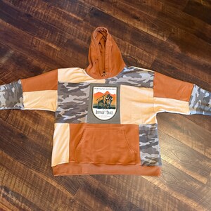 May include: A patchwork hoodie in shades of orange, tan, and camouflage. The hoodie features a kangaroo pocket and a hood. The front of the hoodie has a patch with the text "Buffalo Trace" and an image of a bison. The sleeves are a mix of camouflage and orange.