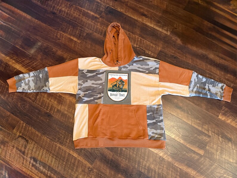 May include: A patchwork hoodie in shades of orange, tan, and camouflage. The hoodie features a kangaroo pocket and a hood. The front of the hoodie has a patch with the text "Buffalo Trace" and an image of a bison. The sleeves are a mix of camouflage and orange.