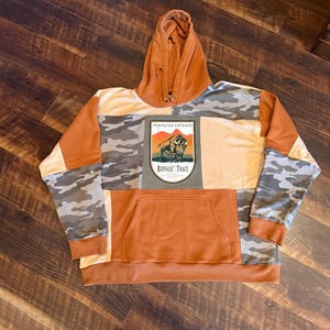 May include: A patchwork hoodie in shades of orange, tan, and camouflage. The front features a kangaroo pocket and a rectangular patch with a bison illustration and the text "Buffalo Trace". The hood is a solid orange color.