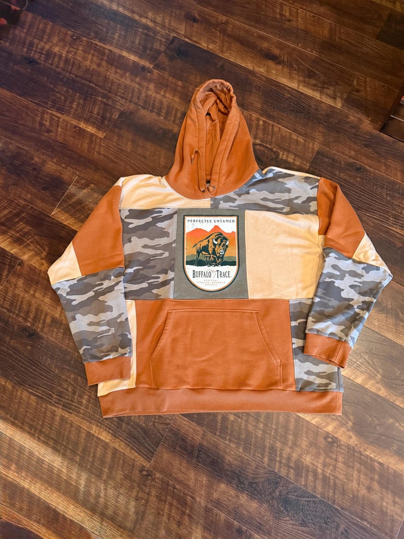 May include: A patchwork hoodie in shades of orange, tan, and camouflage. The front features a kangaroo pocket and a rectangular patch with a bison illustration and the text "Buffalo Trace". The hood is a solid orange color.