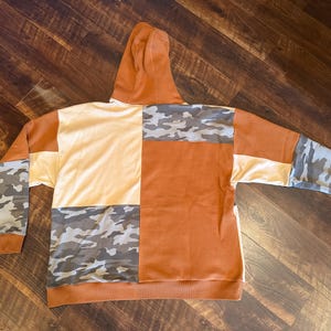 May include: A patchwork hoodie in shades of orange, cream, and camouflage. The hoodie features a hood and long sleeves. The design incorporates blocks of solid colors and camouflage patterns, creating a unique and stylish look. The hoodie is made of a soft material.