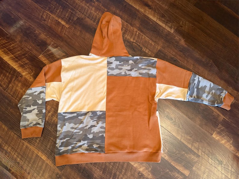 May include: A patchwork hoodie in shades of orange, cream, and camouflage. The hoodie features a hood and long sleeves. The design incorporates blocks of solid colors and camouflage patterns, creating a unique and stylish look. The hoodie is made of a soft material.