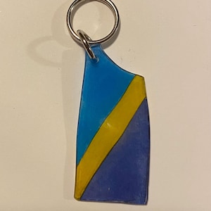 May include: A blue, yellow, and navy blue keychain with a silver key ring. The keychain has a geometric design with a yellow stripe across the center. The top portion is blue, and the bottom is navy blue.