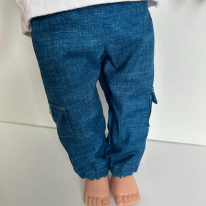May include: A pair of blue cargo pants with a textured fabric finish, featuring side pockets with flaps on each leg, paired with a plain white t-shirt. The pants have a straight-leg cut and are worn by a doll with visible bare feet.