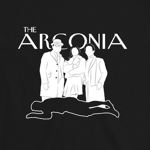 Puede incluir: Camiseta negra con un gr&aacute;fico blanco de tres figuras de pie frente a una figura ca&iacute;da. El texto "THE ARCONIA" est&aacute; por encima de las figuras.