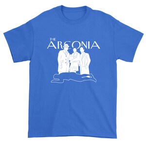 Puede incluir: Una camiseta azul real con un gr&aacute;fico blanco de una familia de cuatro personas de pie frente a la silueta de una persona tendida en el suelo. El texto "THE ARGONIA" est&aacute; por encima de la familia.