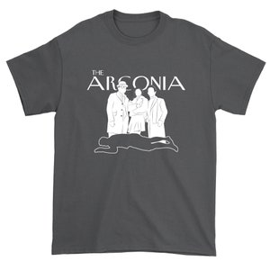 Puede incluir: Una camiseta gris oscuro con un dise&ntilde;o gr&aacute;fico blanco que presenta el texto "THE ARCONIA" y una silueta de tres personas.