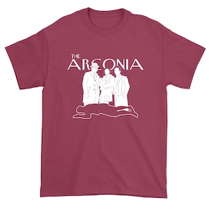 Puede incluir: Una camiseta de color granate con un gr&aacute;fico blanco de tres figuras de pie frente a la silueta de una persona tendida en el suelo. El texto "THE ARCONIA" est&aacute; por encima de las figuras.
