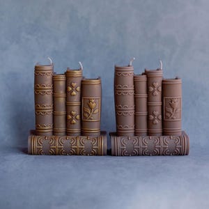 May include: Two sets of brown book-shaped candles with gold accents. Each set has five candles with different designs, including a flower and a clover.