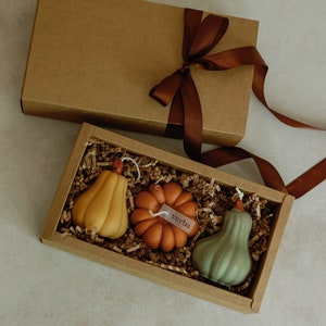 May include: A gift box filled with three small, hand-crafted candles shaped like gourds. The candles are in shades of green, orange, and yellow. The box is brown with a brown ribbon and a small tag that says "verba".
