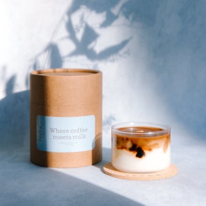 May include: A brown cardboard tube with a blue label that reads "Where coffee meets milk" and a glass of iced coffee with a brown and white layered design. The glass is sitting on a cork coaster.