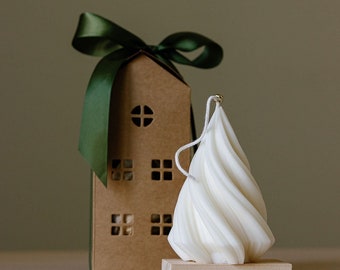 May include: A white, swirled candle with a wick, resting on a small wooden block. A brown house-shaped gift box with cut-out windows and a green ribbon is behind the candle. The candle has a unique, textured design.